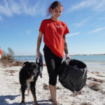 A Petland Pembroke Pines volunteer walks on the shores of a Largo Beach with a trashcan in hand and her Petland dog on her other side.