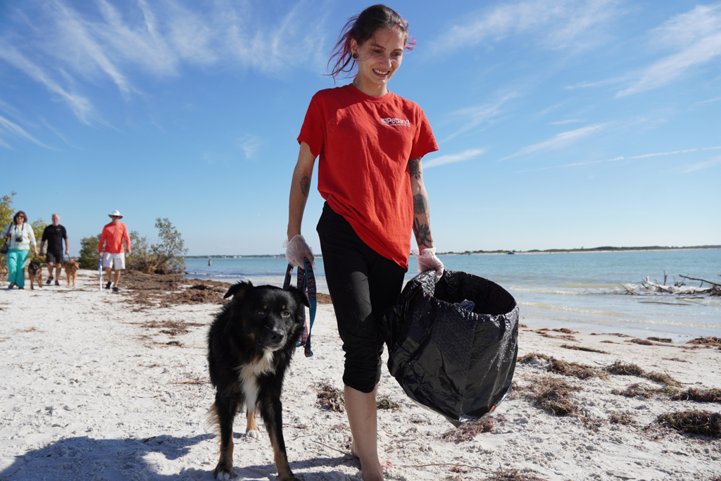 A Petland Pembroke Pines volunteer walks on the shores of a Largo Beach with a trashcan in hand and her Petland dog on her other side.