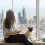 A woman relaxes in the window seat of her city apartment next to her loyal Pug.