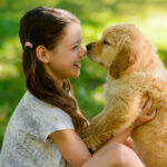 A smiling girl holds a Golden Retriever puppy while sitting in the grass on a sunny day.