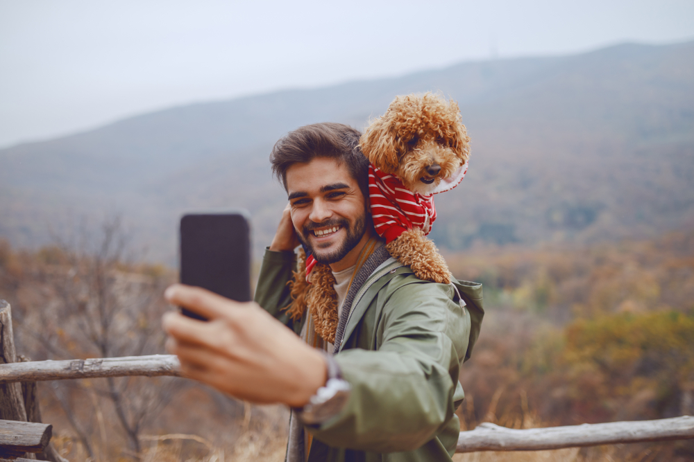 A happy man hikes with his Poodle and takes a selfie with mountains in the background.