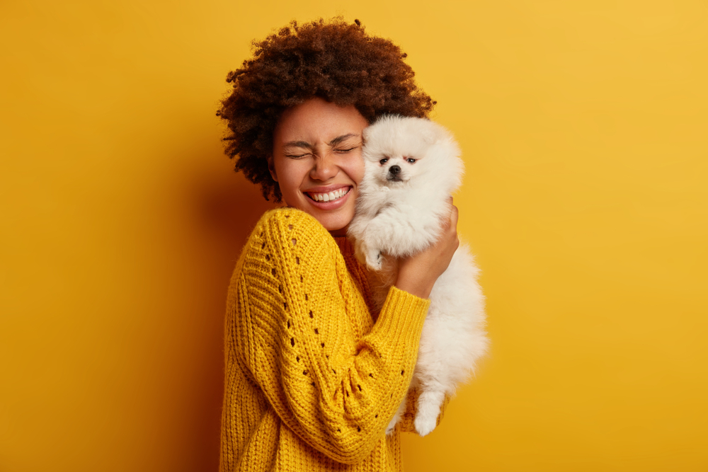 A woman wearing an orange sweater holds a white Pomeranian dog and smiles at how cute her furry friend is!