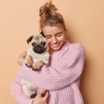 A cozy woman in a pink sweater hugs her cute purebred Pug while standing in front of a peach colored background.