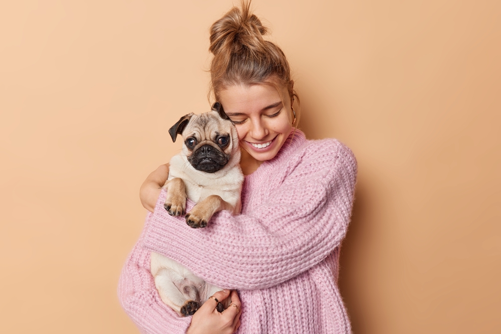 A cozy woman in a pink sweater hugs her cute purebred Pug while standing in front of a peach colored background.