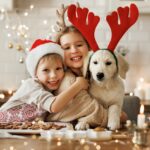 Happy children sit with their yellow Labrador Retriever puppy, all wearing Christmas Santa hats and red reindeer antlers, as they make holiday cookies.